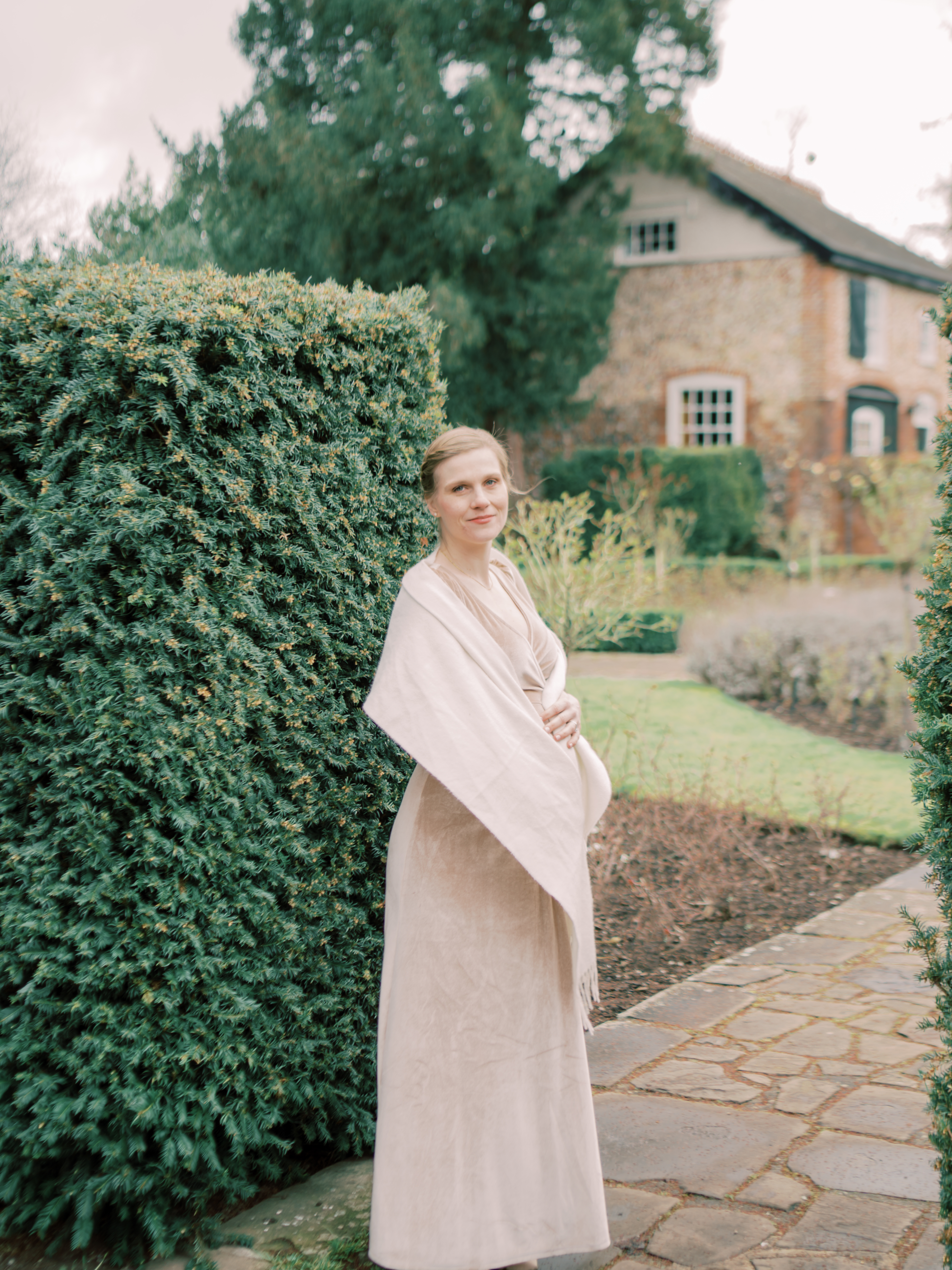 Mother stands in English gardens in front of an English cottage during her professional maternity photo session.