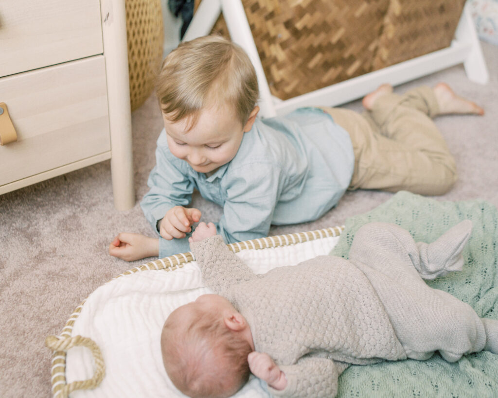 Brother curiously pats his newborn baby sister's hand as she lays in her Moses basket during their family photo session with Oklahoma Newborn Photographer By Courtney Cronin