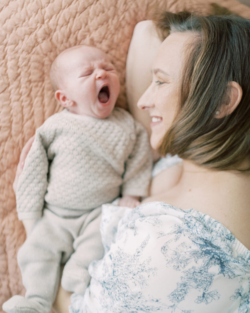 Mother grins at her newborn baby daughter as she yams sleepily during their photo session with By Courtney Cronin
