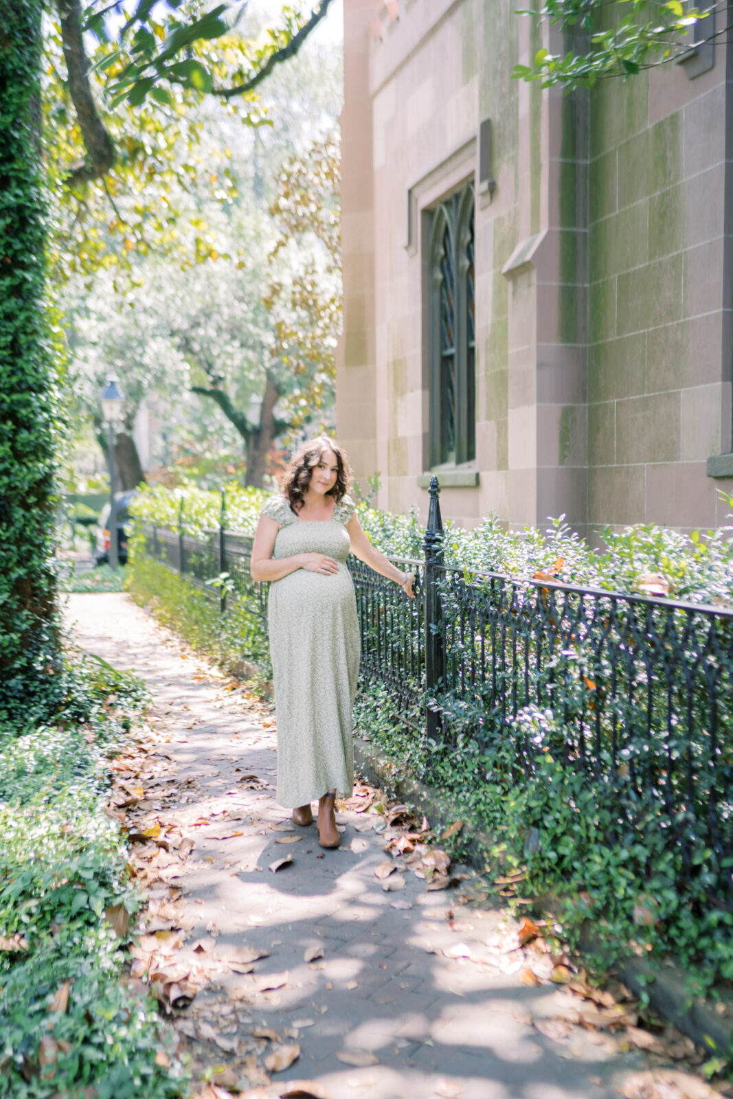 Mother looks at camera while placing her hand on her bump and running her hand along an iron fence in the historic district by Maternity Photographer in Savannah Georgia Courtney Cronin