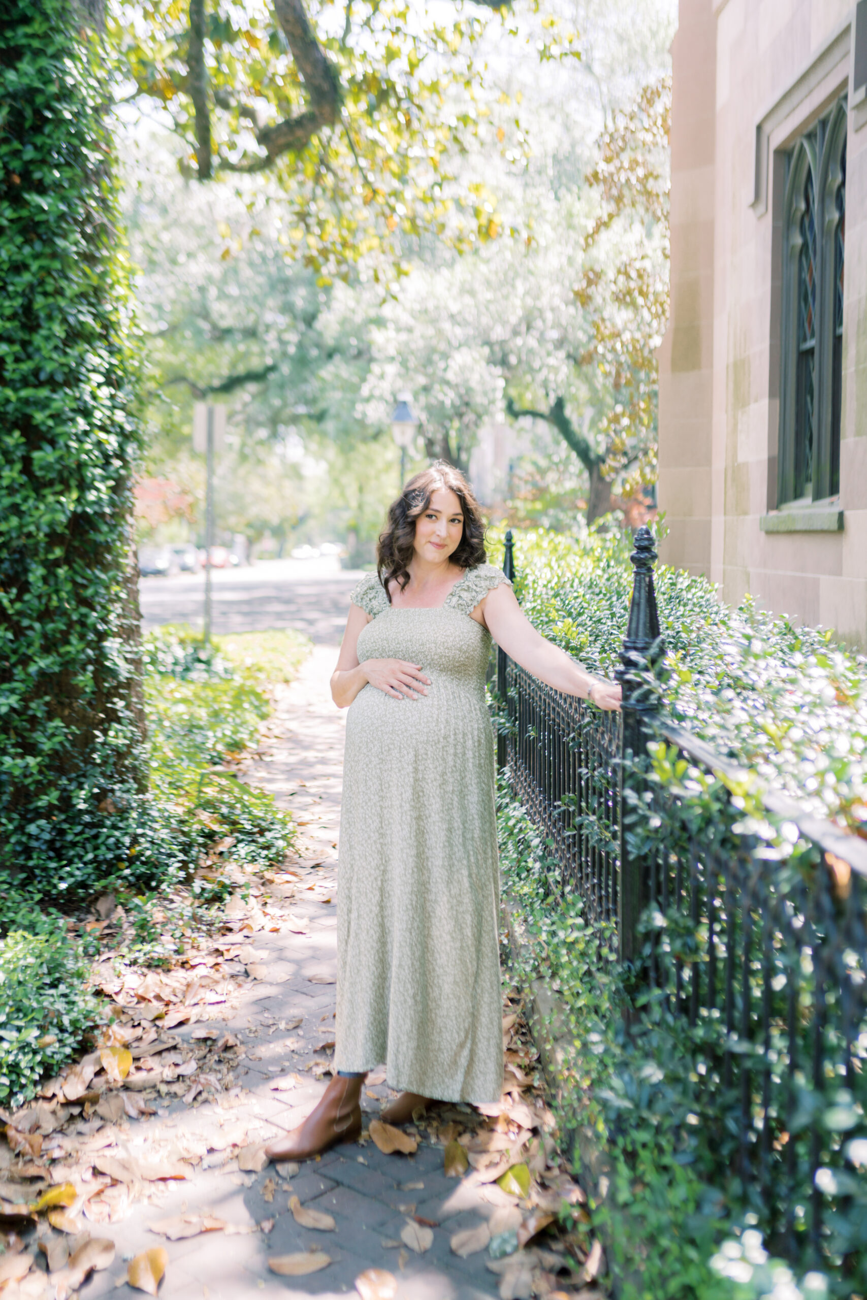 Mother leans on iron fence in downtown historic Savannah and places her hand on her baby bump by Savannah Family Photographer Courtney Cronin
